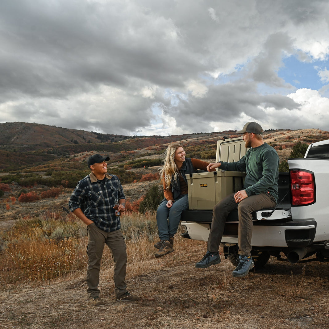 Three people by a white pickup truck in a mountainous area with a cloudy sky.