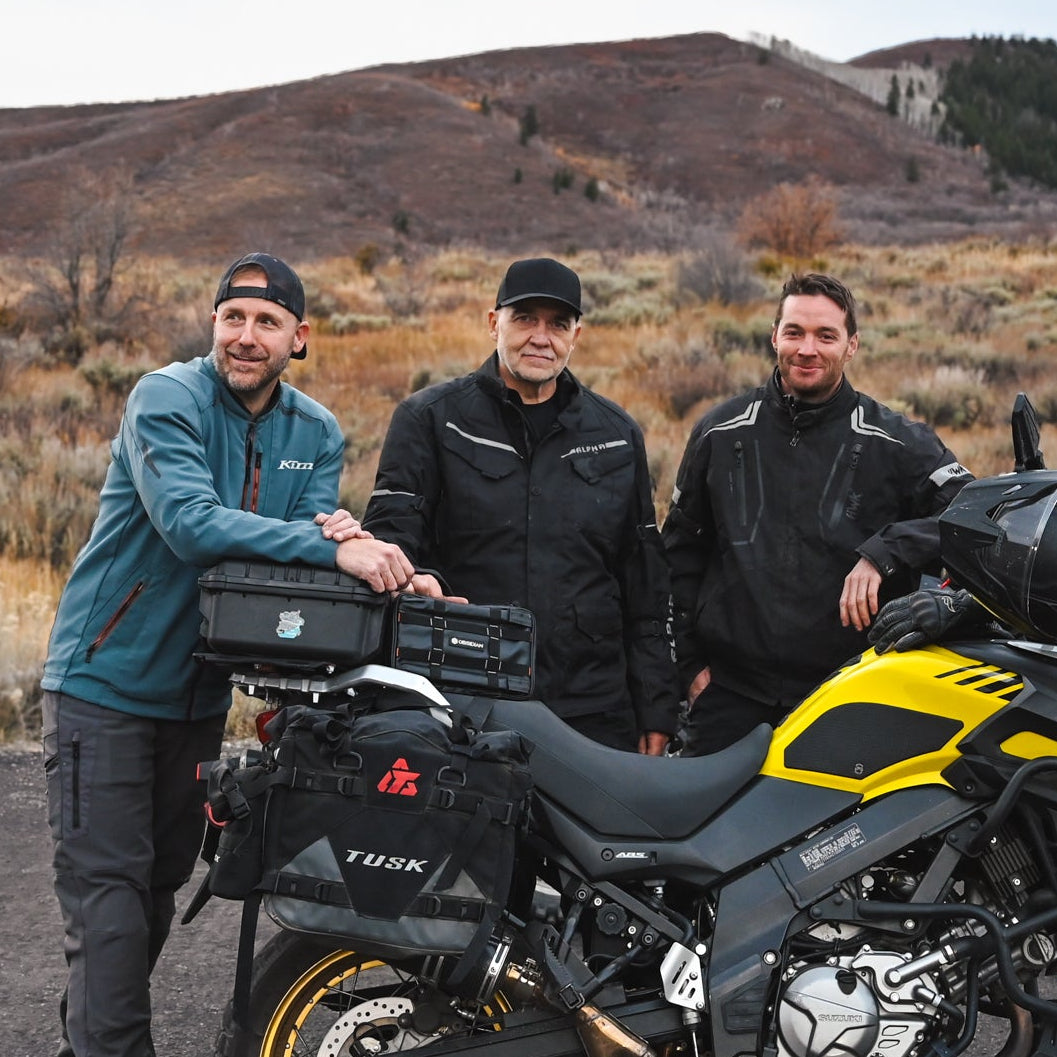 Three people standing next to a motorcycle in an open area with mountains in the background